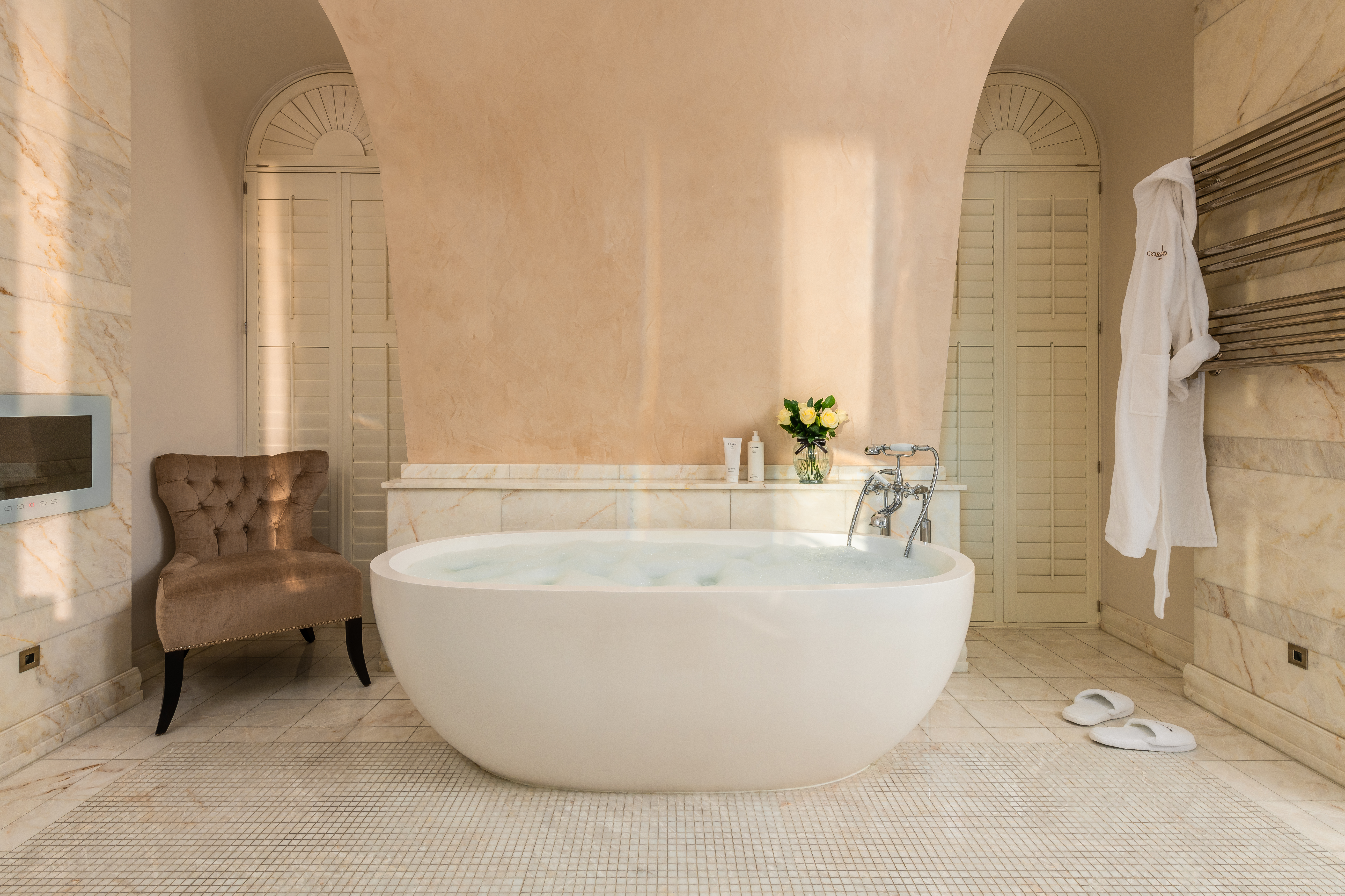Penthouse suite bathroom at Corinthia London luxury hotel featuring large marble tub glass of champagne and robe awaiting guests