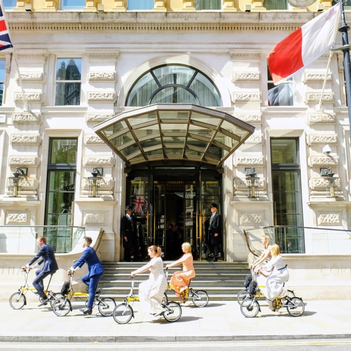 Five people riding Brompton bikes outside hotel entrance