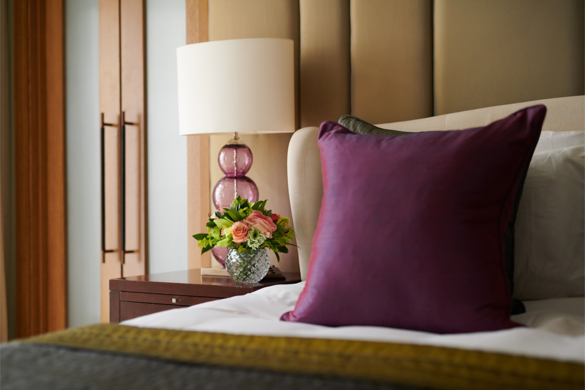 Bed with purple cushion, bedside table with flowers and lamp